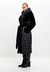 Zwarte lange jas met een faux fur buitenkant en gewatteerde stof, voorzien van een ceintuur in de taille en een hoge kraag, gecombineerd met kniehoge zwarte laarzen.