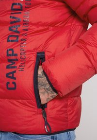 Czerwona kurtka puchowa o strukturze ripstop, granatowe zamki błyskawiczne i kieszeń. Posiada wyraziste logo "CAMP DAVID" w ciemnym kolorze.