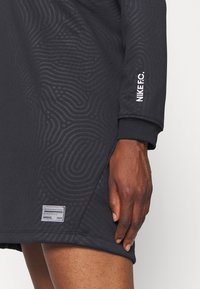 Robe noire Nike FC avec un motif texturé subtil, texte blanc « NIKE F.C. » sur la manche, écusson près de l'ourlet, portée par une personne à la peau brune.