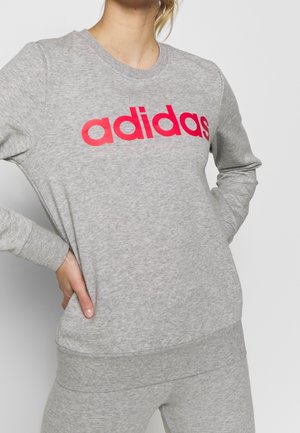 Person iført grå Adidas sweatshirt og matchende bukser med hænderne på hofterne, beskåret fra nakken og ned.