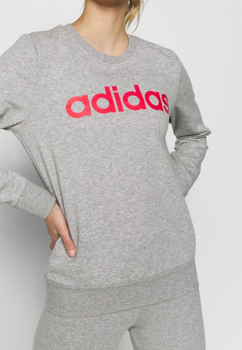 Personne portant un sweat-shirt gris Adidas et un pantalon assorti, les mains sur les hanches, recadrée du cou vers le bas.