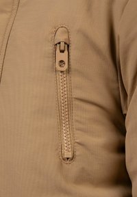 Béžová látka s bundou a detailom so zipsovitou kapsou. Zips má štruktúrovaný úchop a pozdĺž okraja je prešitý.