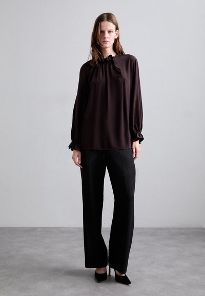 Blusa marrone scuro con scollatura arricciata e maniche lunghe, abbinata a pantaloni neri sartoriali e tacchi neri a punta. Finitura in tessuto liscia.