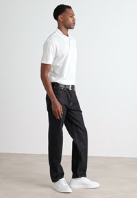 Bílá polokošile, tmavé džínové kalhoty s viditelným švem, černý opasek a bílé tenisky. Model stojící v profilu na neutrálním pozadí.