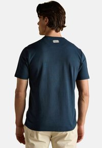 Vīrietis ar muguru pretim, uzvilcis tumši zilu īsroku T-kreklu ar nelielu baltu etiķeti pie kakla un beigu biksēm.