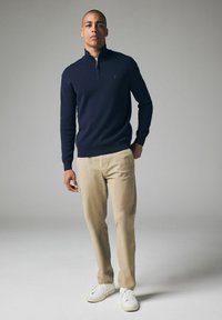Pulover bleumarin cu fermoar, cu mâneci lungi, asociat cu pantaloni bej deschis și adidași albi. Design simplu; textură moale, tăietură slim.
