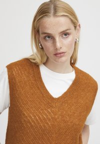 Rostfarbener Strickweste mit V-Ausschnitt und Zickzackmuster, kombiniert mit einem weißen T-Shirt. Glatte Textur und lockere Passform.
