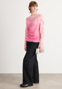 Vrouw in een roze gebreid vest met een patroon en brede zwarte broek, die een kleine zilveren clutch vasthoudt, staand tegen een effen achtergrond.