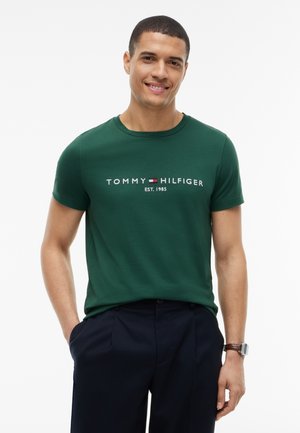 Lächelnder Mann, der ein grünes Tommy Hilfiger T-Shirt und dunkle Hosen trägt, mit den Händen in den Taschen und einer Uhr am linken Handgelenk.
