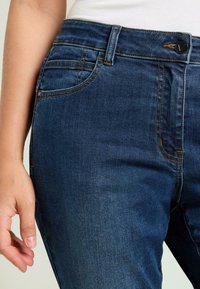 Jeans de mezclilla azul oscuro con un corte ajustado, que presentan costuras contrastantes en naranja, bolsillos delanteros y cierre con botón negro en la cintura.