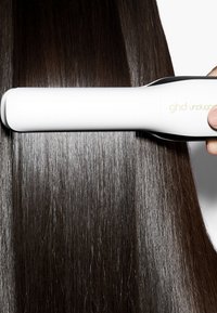 Lisseur à cheveux ghd unplugged blanc, tenu à la main, lissant de longs cheveux bruns foncés et brillants.