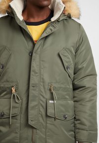 Olivgrüne Parka mit einer mit Pelz gefütterten Kapuze, Druckknopf- und Kordelzugdetails, zwei frontalen Taschen und einem Reißverschluss. Weiche Textur.