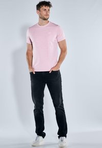Helles rosa T-Shirt mit kurzen Ärmeln und rundem Halsausschnitt, kombiniert mit schwarzen Jeans und weißen Sneakers. Das Model steht vor einem hellen Hintergrund.