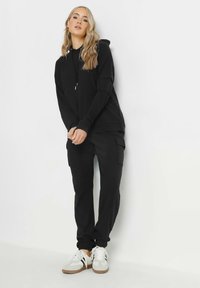 Felpa nera con cappuccio e tasca a marsupio, abbinata a pantaloni cargo neri con tasche laterali. Indossata con sneakers bianche.