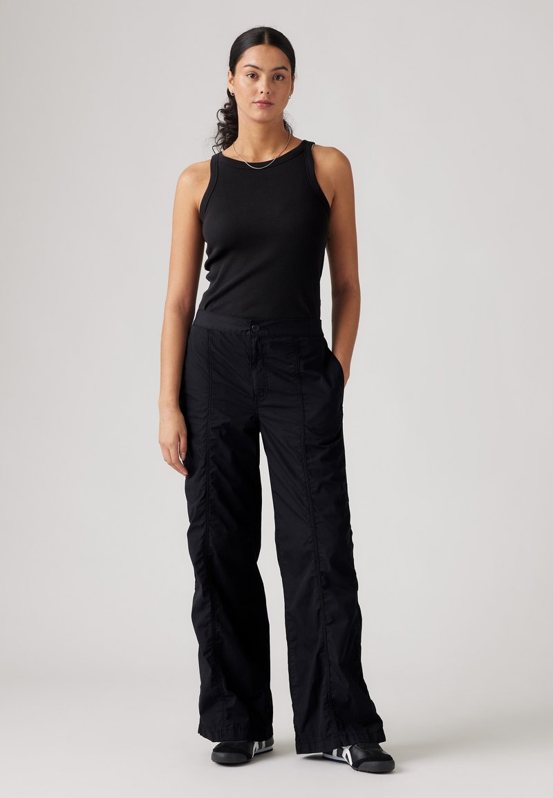 Schwarzes geripptes Tanktop kombiniert mit lockeren schwarzen Stoffhosen mit Knopfverschluss und seitlichen Taschen. Einfaches Design und matte Oberfläche.