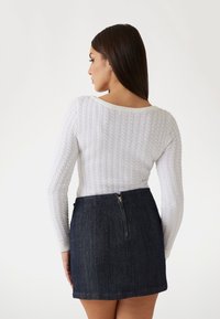 Biela sveter s pletenou textúrou a dlhými rukávmi, kombinovaný s tmavomodrou denimovou mini sukňou s detailom na zips na zadnej strane. Textúry dobre kontrastujú.
