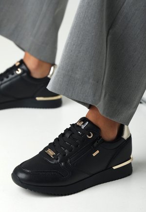 Black leather sneakers with gold accents, round toe, rubber sole, and textured detailing. Paired with light grey tailored pants.