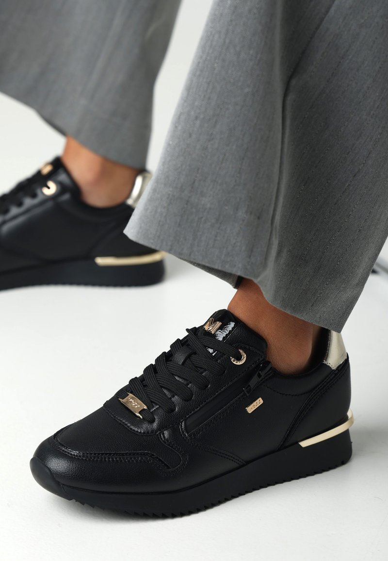Černé kožené tenisky se zlatými akcenty, kulatou špičkou, gumovou podrážkou a texturovanými detaily. V kombinaci se světle šedými kalhotami na míru.