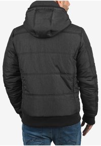 Svart isolert jakke med hette, med teksturert stoff, ribbestrikkede mansjetter og kontrasterende sømmer. Bak har den horisontale paneler.