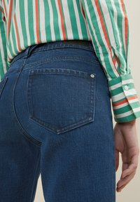 Dunkelblaue Denim-Jeans mit einer Gesäßtasche und einem silbernen Knopf, kombiniert mit einem grün gestreiften Langarmhemd mit roten Akzenten.