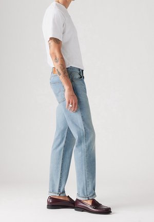 Personne portant un t-shirt blanc, un jean bleu clair et des mocassins en cuir rouge foncé, debout de profil devant un fond uni.