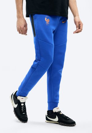 Nedre del av en person som har på seg blå Nike joggebukser med det franske fotballforbundets logo og svarte Nike-sneakers med hvite swoosher.