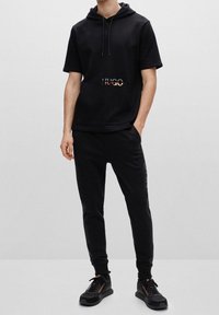 Sort kortærmet hoodie med en frontlomme og et udskæringslogo; parret med sorte sweatpants og matchende sorte sko.