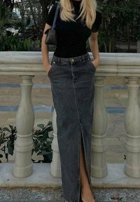 Mujer rubia apoyada en un balaustre de piedra, vestida con una blusa negra, una larga falda de mezclilla oscura con abertura frontal, manos en los bolsillos y llevando un bolso negro de hombro.