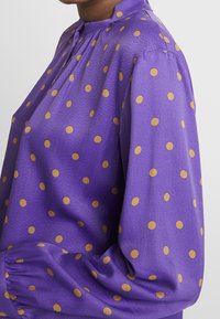 Lila blus tillverkad av ett slätt tyg, prydd med guldprickar och en samlad halsringning. Långa ärmar med elastiska manchetter.
