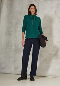 Teal gestrickter Pullover mit Kapuze und Kordeln, kombiniert mit dunkelblauen weit geschnittenen Jeans und schwarzen Stiefeletten. Der Hintergrund ist neutral.