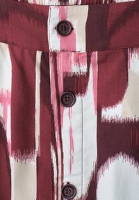 Gros plan sur un tissu à motifs bordeaux, rose et blanc avec trois boutons bordeaux foncé alignés verticalement sur une patte cousue.