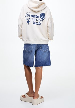 Person facing away, wearing a cream hoodie with text and dice graphic, denim shorts, and beige platform sandals against a plain background.