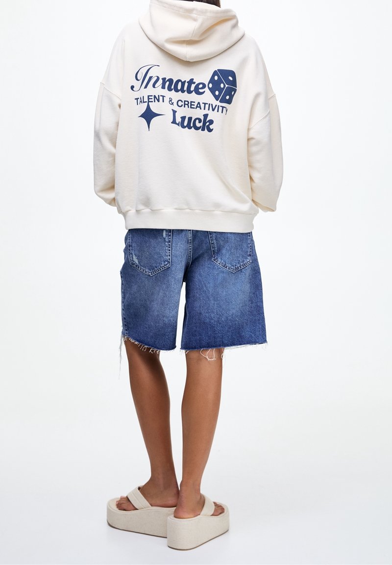 Personne de dos, portant un hoodie crème avec texte et graphique de dés, short en jean et sandales à plateforme beiges sur fond uni.