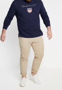 Marineblauses Hemd mit "GANT"-Logo und Wappen-Grafik, kombiniert mit beigen hochgekrempelten Hosen und weißen Sneakern. Lässiges Outfit.