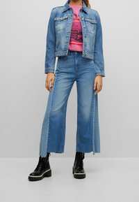 Osoba v džínové jacketě, růžovém tričku s grafikou, širokých dvoubarevných modrých džínách a černých šněrovacích botách stojí na jednobarevném pozadí.