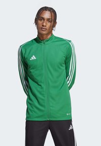 Groene vest met rits gemaakt van een glad materiaal, met drie witte strepen langs de mouwen en een Adidas-logo op de borst.