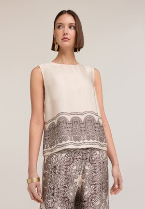 Femme portant un haut beige sans manches et un pantalon large assorti avec des motifs géométriques marron foncé complexes, accessoirisée avec un bracelet et des boucles d’oreilles en or.