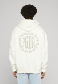 Sudadera blanca con capucha y un gran emblema floral beige que presenta el texto "PGDR" en la espalda. Cuenta con capucha con cordones y un ajuste holgado.