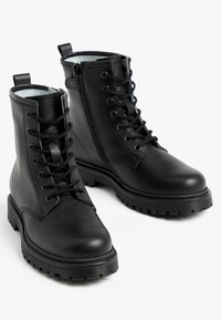 Bottines en cuir noir avec un laçage à l'avant et une fermeture éclair sur le côté. Elles disposent d'une semelle robuste avec un motif texturé pour une meilleure adhérence.