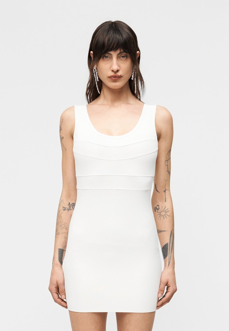 Άτομο με μακριά σκούρα μαλλιά και τατουάζ στα χέρια, που φοράει αμάνικο εφαρμοστό λευκό φόρεμα και μακριά κρεμαστά σκουλαρίκια, στέκεται μπροστά σε απλό φόντο.