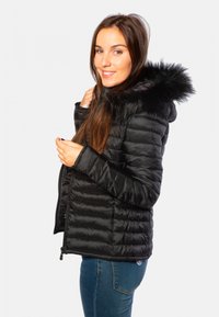 Doudoune noire avec une capuche en fausse fourrure, design matelassé, fermeture éclair avant et coupe ajustée. Associée à un jean bleu, affichant un look décontracté.