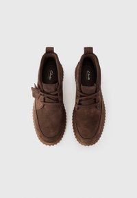 Chaussures décontractées en daim brun avec bouts ronds, lacets texturés et semelles en caoutchouc à rainures saillantes. Logo visible sur la semelle intérieure.