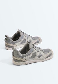 Paire de baskets athlétiques gris et beige avec panneaux en mesh et fermeture à lacets, présentée côte à côte sur fond blanc.