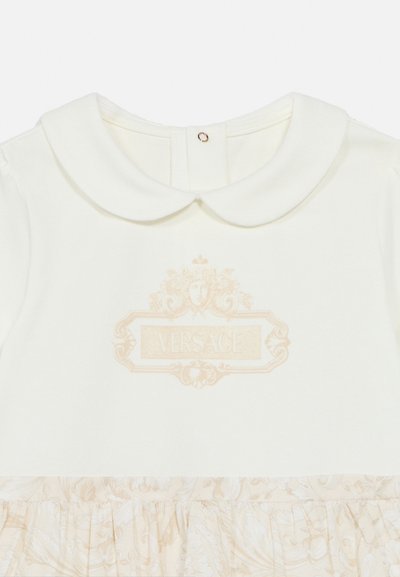 Λευκό βρεφικό φόρεμα με γιακά Πίτερ Παν, φούστα με απαλό λουλουδάτο μοτίβο και μπεζ περίτεχνο λογότυπο Versace στο στήθος.