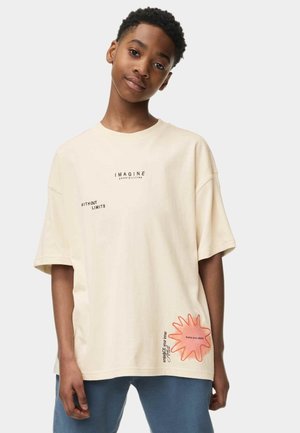 Beige oversized katoenen T-shirt met korte mouwen; heeft de opdruk "IMAGINE" en een roze stervormige grafiek aan de zoom.