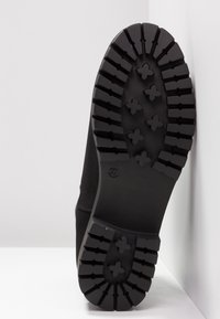 Semelle de chaussure noire avec des crampons profonds en forme de croix, taille 37 embossée près de la voûte plantaire.