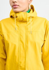 Chaqueta impermeable amarilla con alto collar, cordones ajustables y textura suave. Cuenta con herrajes plateados y un diseño minimalista.