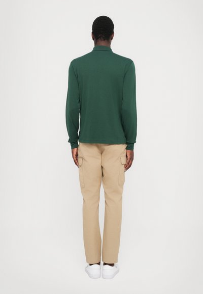Camisa polo de manga larga verde combinada con pantalones cargo beige y zapatillas blancas. Diseño ajustado con textura suave y puños acanalados.