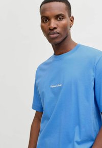 Sinine puuvillane T-särk lühikeste varrukatega, millel on valge "Original Studio" tikand rinnal. Sile tekstuur ja regulaarne lõige.