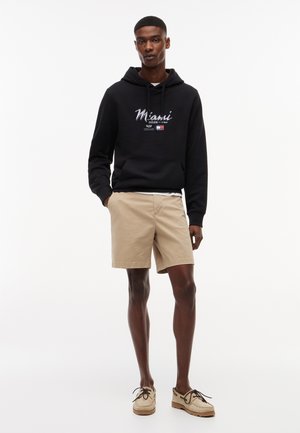 Fiatal férfi, kezét a zsebében, fekete kapucnis pulóverben, melyen "Miami 2026" felirat látható, bézs rövidnadrágban és világosbarna fűzős cipőben, fehér háttér előtt áll.